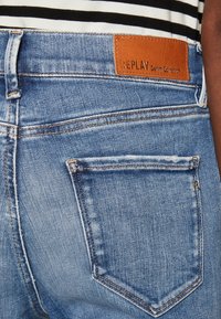 Sinine denimist teksapüksid tekstureeritud kangast, millel on pruun nahkpatch, millel on kirjas "REPLAY Denim Collection" vöökoht.
