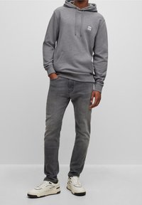 Felpa grigia con cappuccio, dotata di tasca frontale e patch con logo, abbinata a jeans grigi e sneakers bianche con dettagli neri.