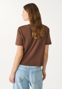 Braunes Kurzarmshirt mit lockerer Passform und rundem Ausschnitt. Kombiniert mit hellblauen, hochtaillierten Jeans, Ansicht von hinten und aus weichem Stoff.