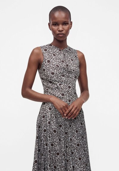 Femme aux cheveux courts portant une robe noire sans manches avec un motif floral de marguerites blanches, debout sur un fond blanc uni.