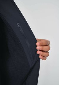 Dunkelblaue Jacke aus glattem Außenmaterial. Verfügt über eine Reißverschlusstasche und eine strukturierte Fütterung. Eine Hand hält die Jacke an der Öffnung.