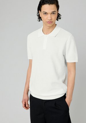 Polo shirt blanc à manches courtes avec une finition texturée, patte de boutonnage à trois boutons et col classique, associé à un short foncé.