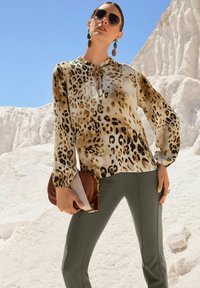 Crème en bruine luipaardprint blouse met lange mouwen, geplooide kraag en trekkoorden. Olijfgroene strakke broek, bruine handtas, zonnebril en statement oorbellen.