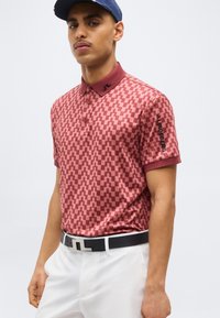 Férfi, piros mintás galléros pólóban, fehér nadrágban, fekete övvel és sötétkék sapkával, egyszínű háttér előtt áll.