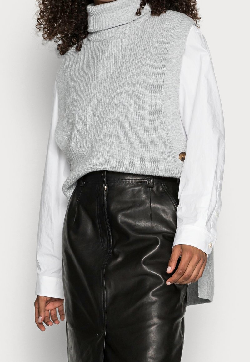 Un pull en maille grise à col roulé superposé sur une chemise blanche à manches longues, associé à une jupe en cuir noir avec fermeture éclair sur le devant.