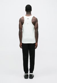 Muž stojaci chrbtom, oblečený v bielom športovom tielku bez rukávov, čiernych nohaviciach, bielych ponožkách a čiernych topánkach na jednoduchom pozadí.