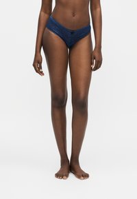 Γυναίκα που στέκεται ξυπόλητη σε λευκό φόντο, φοράει σκούρα μπλε δαντελένιο εσώρουχο, εμφανίζεται από τη μέση μέχρι τα πόδια.