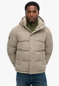 Kurtka puchowa w jasnobeżowym kolorze, z kapturem, zapięciem na zatrzaski, pikowanym wzorem i bocznymi kieszeniami, noszona na zielonym swetrze z dzianiny.