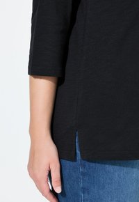 Haut noir texturé à manches trois-quarts, avec une fente sur le côté et associé à un denim bleu. Détails de couture subtils sur les manches.