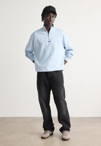 Sudadera de cremallera de color azul claro con cuello levantado y puños acanalados, combinada con jeans oscuros y zapatillas grises. Branding mínimo en la sudadera.