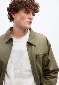 Νέος άνδρας με σγουρά μαλλιά που φοράει ένα χακί πράσινο μπουφάν πάνω από ένα λευκό γραφικό T-shirt με σχεδίαση δακτυλίων δέντρου και το κείμενο «BOSS».