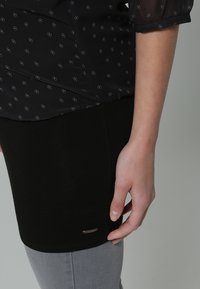 Buste latéral d'une femme avec un chemisier noir à motifs, une jupe noire et un pantalon gris, main reposant naturellement le long du corps sur un fond gris.