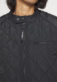 Gros plan sur une veste matelassée noire avec une fermeture éclair et un col à boutons-pression, comportant une poche zippée horizontale sur la poitrine.