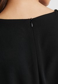 Svart klädtyg med en slät textur. Har en bakstängning med en liten svart dragkedja, placerad vid halsringningen.