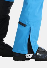 Blaue isolierte Skihosen mit schwarzen Akzenten, Reißverschlussöffnungen an den Bündchen und einem Markenlogo. Skischuh teilweise sichtbar.