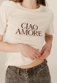 T-shirt en coton crème à manches courtes, avec "CIAO AMORE" imprimé en marron foncé. Associé à un jean gris à imprimé léopard.
