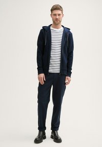 Hanorac cu fermoar de culoare bleumarin peste un tricou alb cu dungi bleumarine, asortat cu pantaloni bleumarin și cizme negre până la gleznă. Textură fină și croială lejeră.