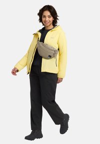 Giacca impermeabile gialla, base layer nera, pantaloni neri e stivali neri. Indossa una borsa a tracolla beige con zip.