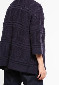 Marinblå stickad cardigan med strukturerade geometriska mönster, trekvartsärmar och lös passform, med öppenhet detaljer vid axlarna.