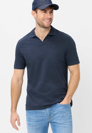 Lächelnder Mann, der ein marineblaues Poloshirt, hellblaue Jeans und eine dunkelblaue Kappe trägt, steht mit einer Hand in der Tasche vor einem schlichten Hintergrund.