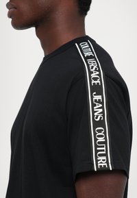 Προφίλ από το πλάι ενός ατόμου που φοράει ένα μαύρο T-shirt με λωρίδα κειμένου "Couture Versace Jeans Couture" που εκτείνεται από τον ώμο μέχρι την άκρη του μανικιού.