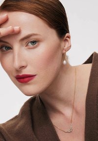 Złoty naszyjnik o zakrzywionym wzorze z małymi przezroczystymi kamieniami szlachetnymi. Kolczyki w kształcie kropli z perłami. Modelka ma na sobie brązowy sweter. Neutralne tło.