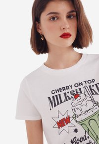 Witte katoenen t-shirt met een grafische print van een milkshake met een kers, accentueert in groen en rood. Ronde hals en korte mouwen.
