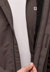 Bruine zip-up jas met een gestructureerde buitenkant en zachte fleece voering, gedeeltelijk open. Een lichtgrijze shirt is zichtbaar eronder.