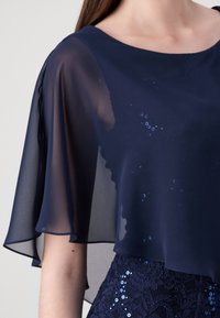 Žena v námořnickém modrém šatu s průhlednými, splývavými rukávy a korzetem pokrytým flitry a krajkou.