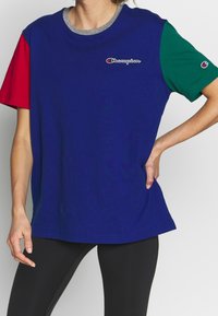 Žena v modrém tričku Champion s červeným levým rukávem, zeleným pravým rukávem, šedým límcem a černými legínami, stojící s rukou na boku.