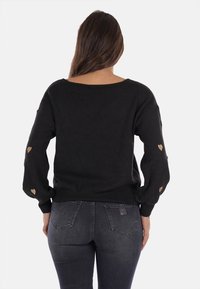 Maglione nero a maniche lunghe, con dettagli a forma di cuore dorato sulle maniche. Il capo ha una vestibilità morbida e un ampio scollo.