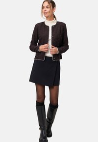 Zwarte textuurjacket met zilveren details, ronde halslijn en knoopsluiting. Draagt over een crèmekleurige coltrui, gecombineerd met een zwarte rok en kniehoge laarzen.