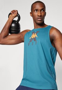 Mann som løfter en svart kettlebell med høyre arm, iført en turkise ermeløs treningsskjorte med teksten "K FIT", stående mot en ensfarget bakgrunn.