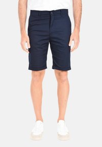 Pantaloni corti in cotone blu navy con chiusura a bottone, risvolti e tasche frontali. Indossati con sneakers bianche su uno sfondo neutro.