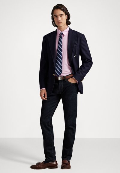 Blazer navy, camicia rosa chiaro, cravatta a righe, jeans in denim scuro, mocassini in pelle marrone. Il blazer presenta accenti in hardware dorato e dettagli sulle tasche.
