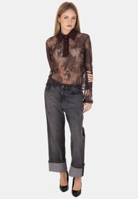 Giovane donna che indossa una blusa in pizzo nero trasparente con colletto e polsini scuri, abbinata a jeans grigi larghi con risvolti e tacchi neri.