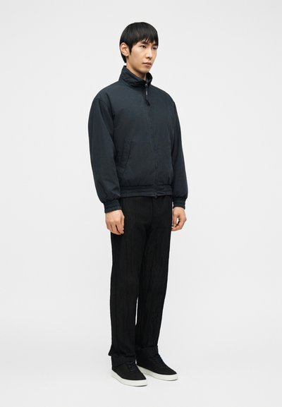 Άνδρας που στέκεται φορώντας μαύρο μπουφάν με φερμουάρ, μαύρο παντελόνι και μαύρα αθλητικά παπούτσια με λευκές σόλες, μπροστά σε απλό γκρι φόντο.