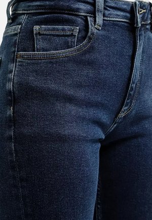 Granatowe dżinsy o gładkiej strukturze, z standardowymi kieszeniami, szlufkami na pasek i kontrastującymi szwami wzdłuż krawędzi.