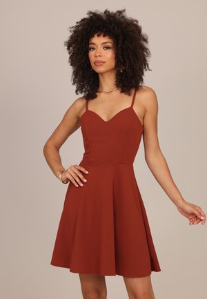 Donna con capelli ricci che indossa un vestito smanicato color ruggine con sottili spalline, posando con una mano sul fianco e l'altra leggermente estesa.