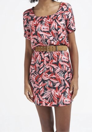 Kobieta ubrana w sukienkę z krótkim rękawem w różowo-czerwony wzór liści, z brązowym paskiem z ćwiekami w talii.