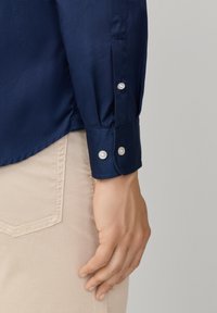 Chemise bleu marine avec une texture lisse, dotée d'un poignet à boutons et de trois boutons en coquillage sur la manche, associée à un pantalon beige clair.