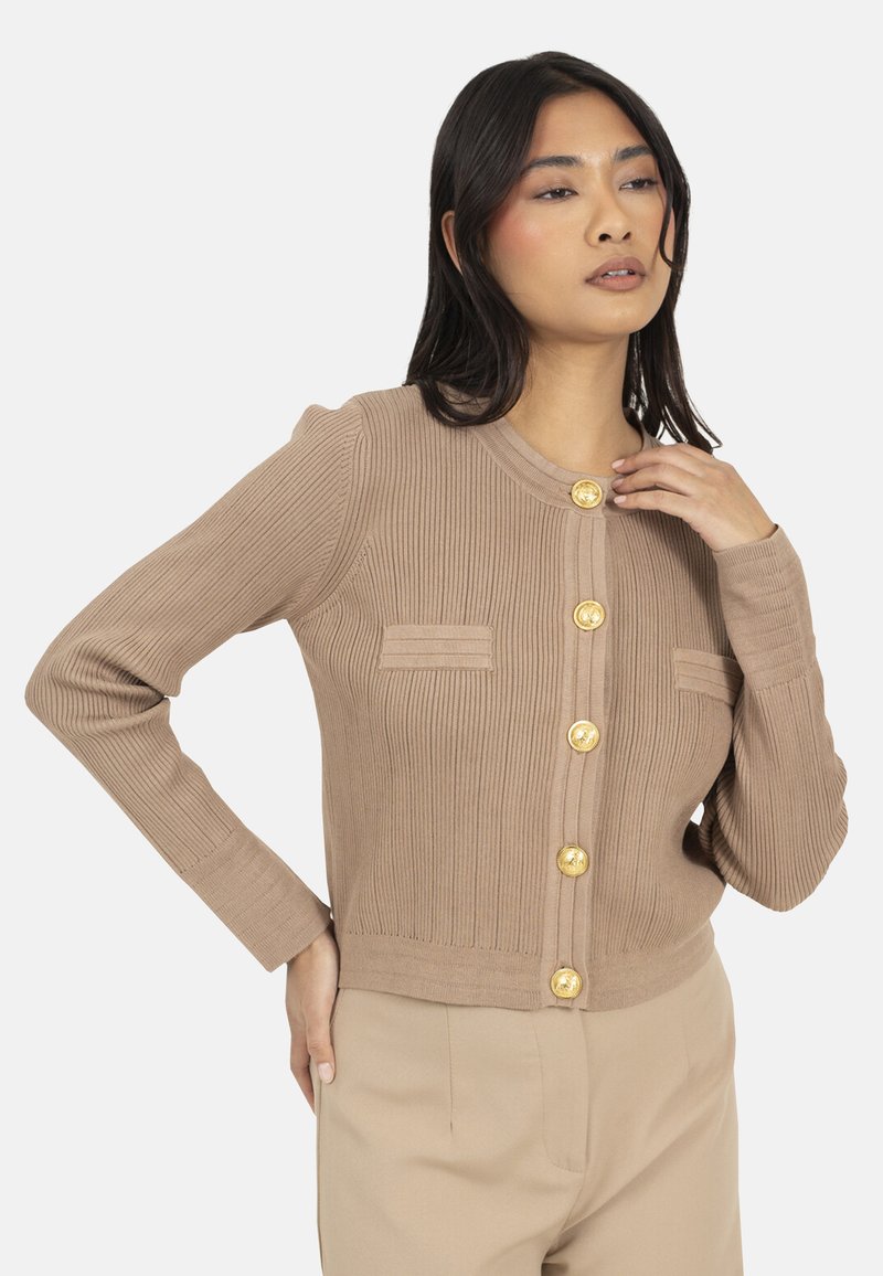 Cardigan beige à côtes avec de longues manches, boutons dorés et deux petites poches sur la poitrine. Tissu doux et design ajusté.