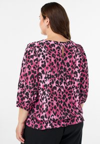 Ženska, oblečena v bluzo s potiskom leoparda v roza in črni barvi, z rokavi do tri četrtine in majhnim gumbom s ključavnico na zadnjem izrezu vratu.