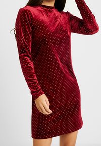 Röd sammetsklänning med långa ärmar, prickigt mönster, hög halsringning och en slim fit-design. Slät textur med en lätt glans.