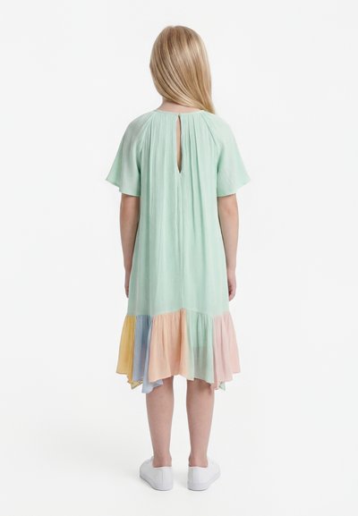 Fiatal lány hosszú szőke hajjal, mentazöld ruhában, amelynek színes fodros alja van, és fehér tornacipőt visel, háttal áll.