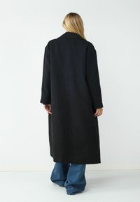 Lange zwarte jas met een gestructureerde afwerking, brede mouwen en een klassieke kraag. Gecombineerd met uitlopende blauwe denim broek.