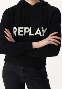 Schwarzer, gerippter Kapuzenpullover aus weichem Material mit einem auffälligen weißen "REPLAY"-Logo über der Brust. Das Design umfasst eine Kapuze mit Kordelzug.