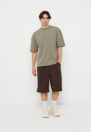 Junger Mann, der ein übergroßes olivgrünes T-Shirt, dunkelbraune knielange Shorts, weiße Socken und beige Turnschuhe trägt, steht vor einem einfarbigen weißen Hintergrund.