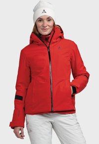Rote wasserdichte Jacke mit Kapuze, schwarzen Akzenten, Reißverschlüssen und Logo. Kombiniert mit weißen isolierten Hosen. Leicht und mit glatter Textur.