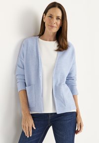 Vaaleansininen ribattu neuletakki, jossa on avointa etuosaa ja kaksi taskua, kerrottu valkoisen paidan päällä. Yhdistetty tummiin farkkuihin, jotka ovat sileäpintaiset.